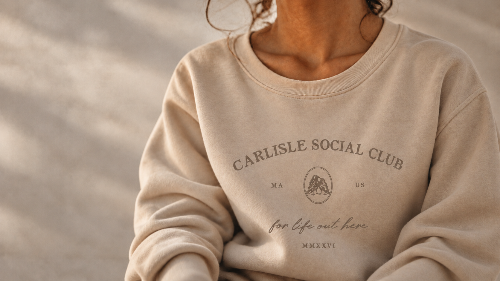 Warm film-toned close-up of a Sand crew sweatshirt with the Carlisle Social Club crow crest