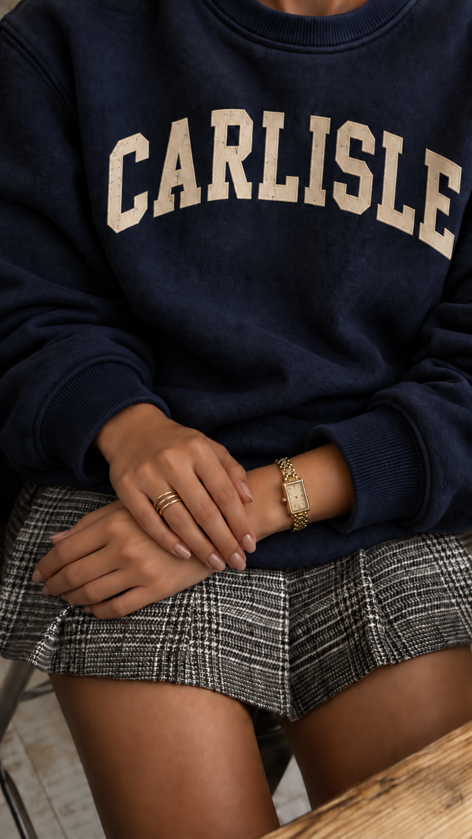 Faceless seated model wearing the Navy Carlisle Collegiate Crew with both hands visible, a gold rectangular watch and gold stacking rings